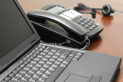 A black laptop computer in the foreground with a slightly out-of-focus business telephone and operator headset in the background.